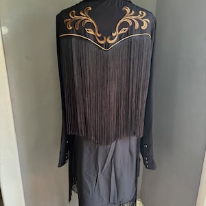 Op de afbeelding: Zwart franjeshirt met gouden borduurwerk op de rug. Het shirt heeft een kraag en lange mouwen.