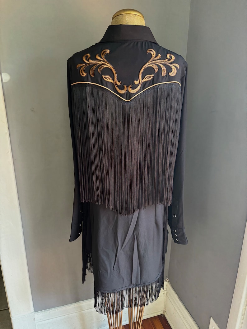 Op de afbeelding: Zwart franjeshirt met gouden borduurwerk op de rug. Het shirt heeft een kraag en lange mouwen.