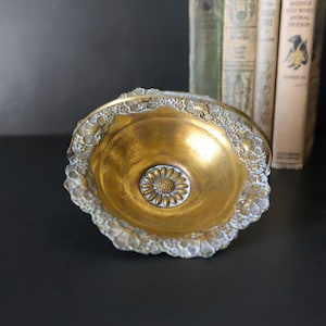 May include: A vintage brass bowl with a floral design in the center and a decorative, ornate edge. The bowl has a golden hue and sits on a dark surface. The bowl is likely used for decorative purposes.
