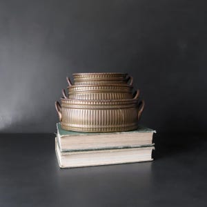 May include: A stack of three round, metallic bronze-toned containers with a ribbed, vertical texture and small side handles. The containers are nested together, decreasing in size from the bottom to the top, and rest upon two stacked hardcover books.