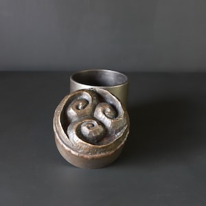 May include: A silver metal container with a lid featuring a three-dimensional design of three swirling shapes.