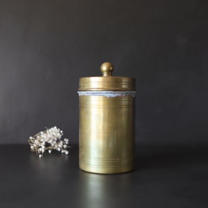 May include: A gold metal container with a lid. The container has a smooth, shiny surface and a slightly worn look. It is sitting on a black surface.