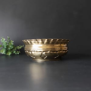 May include: A round brass bowl with a metallic gold finish. The bowl features a scalloped, wavy rim and a decorative embossed pattern around the lower section, consisting of repeating petal-like shapes that provide a textured appearance.