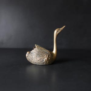 May include: A brass swan figurine featuring a long, slender neck and a body with detailed, etched feather patterns. The metal has a warm, golden-brown tone with a polished finish, showcasing the intricate texture of the wings and tail feathers.
