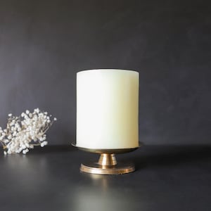 May include: A white pillar candle sits on a gold metal candle holder. The candle holder has a round base and a wide, flat top.