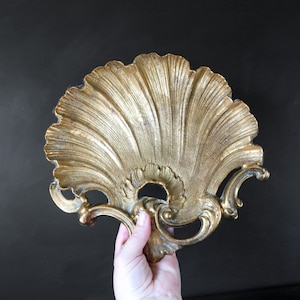 May include: A gold-toned metal decorative tray shaped like a seashell with intricate details. The tray has a fan-like design with a scalloped edge.