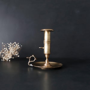 May include: A brass candlestick holder with a round base and a tall, cylindrical body. The candlestick has a handle and a small, round platform at the top. The candlestick is set against a dark background.