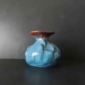 May include: A handcrafted ceramic vase with a unique shape and a glossy finish. The vase features a vibrant turquoise body with a textured surface and a contrasting brown rim. This decorative piece adds a touch of artistry to any space.