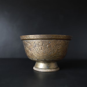 May include: A small, antique brass bowl with intricate floral and animal carvings. The bowl has a wide, rounded base and a flared lip.