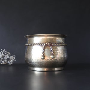 May include: A hammered brass planter with a rope design around the top. The rope has two decorative knots with brass balls at the ends.