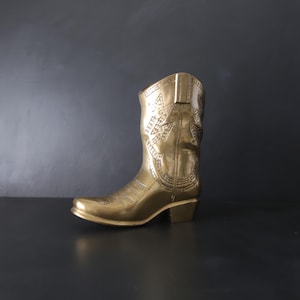 May include: A gold-colored cowboy boot with intricate embossed designs. The boot features a pointed toe, a mid-height heel, and detailed patterns on the shaft. The metallic finish gives it a vintage, decorative appearance.