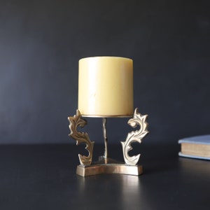 May include: A brass candle holder with a decorative, ornate design. The holder features a star-shaped base and two stylized, leaf-like arms that curve upwards. A yellow candle sits on top of the holder.