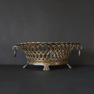 Puede incluir: Una cesta ovalada de latón antiguo con un diseño calado decorativo. La cesta tiene un perfil bajo, con un borde festoneado y pequeños pies. Cuenta con asas de anillo en cada extremo. El latón tiene una pátina cálida y envejecida.
