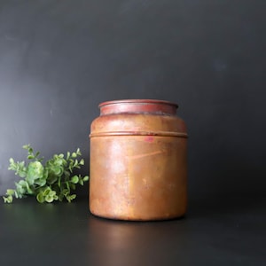 May include: A vintage copper canister with a reddish-brown rim. The cylindrical container has a slightly aged appearance, with visible patina and a warm, metallic hue. The canister is set against a dark background, with a small green plant to the left.