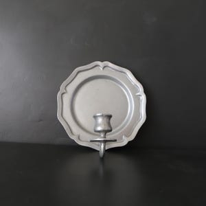 May include: A silver-colored wall-mounted candle holder. The holder features a scalloped plate design with a central candle cup. The metal has a matte finish. The candle holder is designed to be mounted on a wall.
