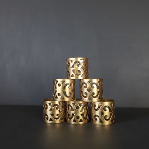 May include: A set of six gold-toned metal napkin rings with an intricate, ornate design. The rings are stacked in a pyramid shape.