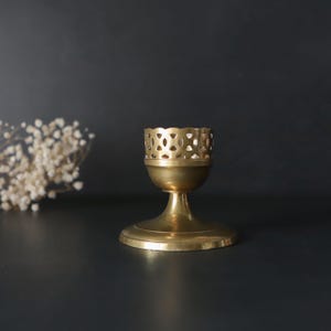 May include: A brass candle holder with a decorative, openwork top and a flared base. The candle holder has a classic, elegant design and is made of shiny, golden-colored metal. The background is a dark, neutral color.
