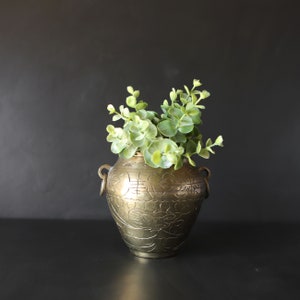 May include: A gold metal vase with an ornate design and two handles. The vase is filled with green leafy plants.