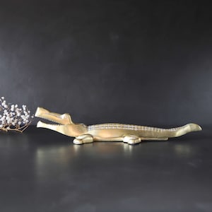 May include: A gold-colored alligator nutcracker with an open mouth and a textured body. The nutcracker is lying on a black surface.