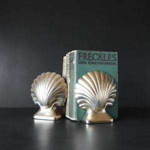 May include: Two silver-colored seashell bookends flank a vintage book titled "FRECKLES" by Gene Stratton-Porter. The bookends have a detailed, fan-like design. The book has a teal cover and is set against a dark background.