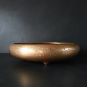 May include: A gold-colored metal bowl with a decorative design of waves and clouds. The bowl has a rounded shape and sits on three small feet.