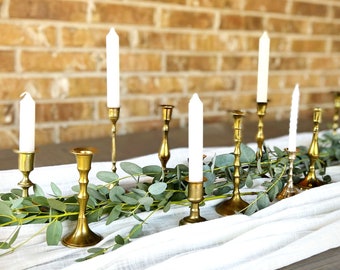 Puede incluir: Un juego de candelabros dorados con velas blancas. Los candelabros están dispuestos sobre un mantel blanco con follaje verde.