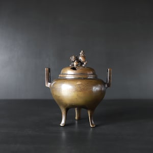 May include: A bronze incense burner with three legs and a lid featuring a decorative animal finial. The burner has a floral design etched into the side.