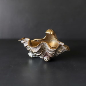 May include: A decorative bowl crafted from brass, shaped like a large clam shell with a textured, rippled exterior and a smooth, polished interior. The object features a metallic gold finish with dark patina accents and rests on small, shell-like feet.