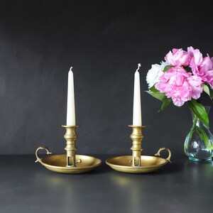 May include: Two gold candle holders with white candles. The candle holders have a round base and a decorative handle. The candles are lit and the holders are on a black surface.