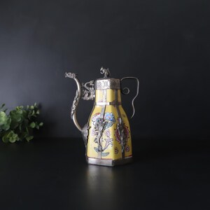 May include: A vintage yellow teapot with floral designs and silver accents. The teapot features a dragon-shaped spout and handle, with a small animal figure on the lid. The body of the teapot is decorated with colorful flowers.