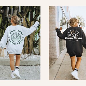 Op de afbeelding: Twee vrouwen dragen zwarte shorts en witte sneakers en dragen sweatshirts met een kampthema. De trui aan de linkerkant is wit met groene tekst en een groen bergontwerp. De trui aan de rechterkant is zwart met witte tekst en een wit bergontwerp. De tekst op de linker trui zegt "Camp Jenna Cabin Crew" en de tekst op de rechter trui zegt "Greetings from Camp Jenna Lake Tahoe".