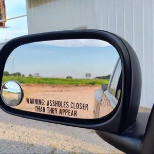 以下が含まれることがあります： 「WARNING: ASSHOLES CLOSER THAN THEY APPEAR」のテキストが入った、黒い車のサイドミラー。 ミラーは田園風景を映し出し、より広い視野を得るための小さな凸面鏡が付いています。