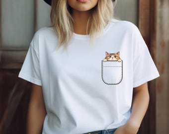 May include: White t-shirt featuring a graphic of a cat peeking out of a pocket. The cat has brown and tan fur with a white chest and paws. The pocket is outlined in black stitching.