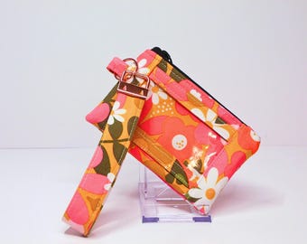 May include: A floral print wallet and wristlet set. The wallet features a clear window and a black zipper. The wristlet strap has a rose gold clasp. The pattern includes orange, pink, white, and olive green flowers.