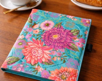 May include: A quilted notebook cover with a vibrant floral pattern. The cover features large pink and orange flowers on a teal background. A cup of coffee and a pastry are in the background.
