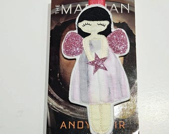 May include: A handmade fabric bookmark featuring a whimsical angel design. The angel has black hair, closed eyes, and glittery pink wings. She holds a pink star. The bookmark is on a copy of "The Martian" by Andy Weir.