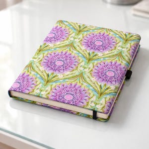 May include: A closed notebook with a floral pattern featuring large purple flowers with pink centers on a light green and blue background. The notebook has a black elastic band closure and is on a white surface.