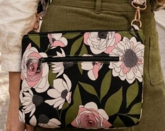 May include: Black crossbody bag with a floral pattern featuring pink and white flowers and green leaves. The bag has a black zipper and a matching floral strap with rose gold hardware. The bag is made of a durable fabric.