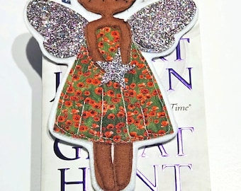 May include: A handmade fairy bookmark with a black bun hairstyle, brown skin, and a green dress with red floral patterns. The fairy has glittery silver wings and is placed on a book. The bookmark is a unique accessory for book lovers.