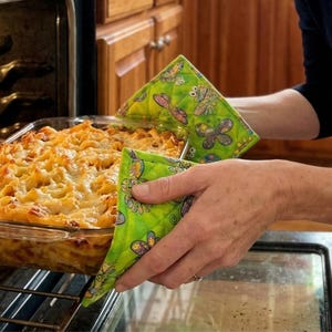 以下が含まれることがあります： オーブンからパスタの入ったガラス製のベーキングディッシュを取り出すために、蝶と花の模様が入った緑色のキルトオーブンミトンを使用している人。ミトンはキルトデザインのライムグリーンです。