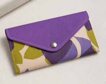 May include: A rectangular fabric wallet with a purple flap secured by a silver snap. The wallet's body features a colorful pattern of green, purple, and beige abstract shapes. The wallet is closed.