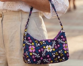 May include: A dark purple shoulder bag with a floral pattern in pink, white, yellow, and blue. The bag has a curved shape and a purple zipper. The strap and the bag's exterior feature the same floral design.