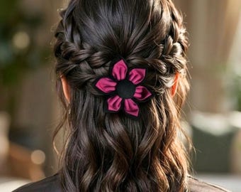 May include: A woman's dark brown hair styled in a braid and wavy style, adorned with a magenta flower hair clip. The flower has a black center and magenta petals. The hair accessory adds a touch of elegance to the hairstyle.