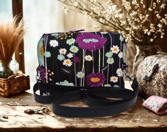 May include: A black crossbody bag with a floral pattern featuring purple and white flowers. The bag has a black adjustable strap and a zipper closure. The bag is sitting on a wooden surface.