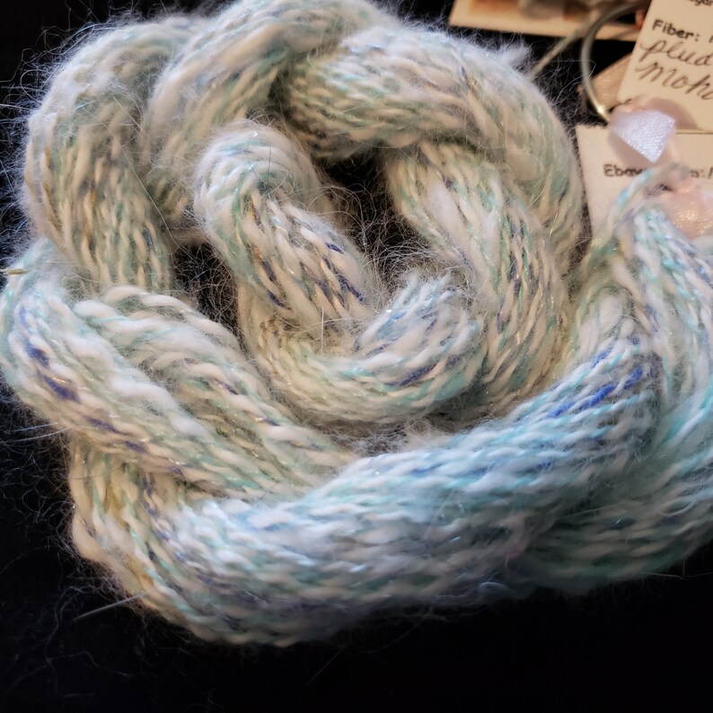 May include: A skein of yarn in shades of white, blue, and green with a subtle shimmer. The yarn is soft and fluffy, and it is perfect for knitting or crocheting.