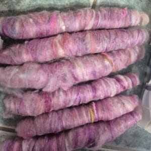 May include: A collection of six thick, hand-spun yarn skeins arranged in a horizontal stack. The yarn features a variegated blend of soft pink, deep magenta, white, and subtle orange fibers with a fuzzy, textured appearance and occasional metallic shimmer.