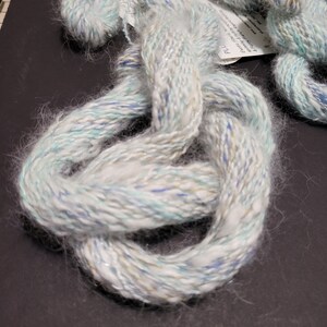 May include: A skein of white yarn with blue, green, and gold flecks. The yarn is soft and fluffy, and it has a subtle shimmer.