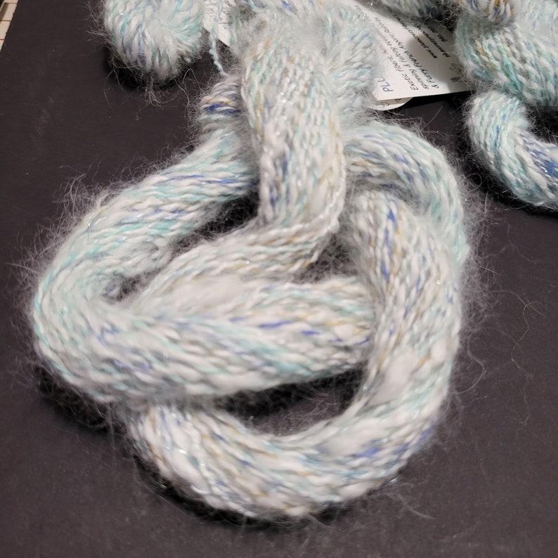 May include: A skein of white yarn with blue, green, and gold flecks. The yarn is soft and fluffy, and it has a subtle shimmer.