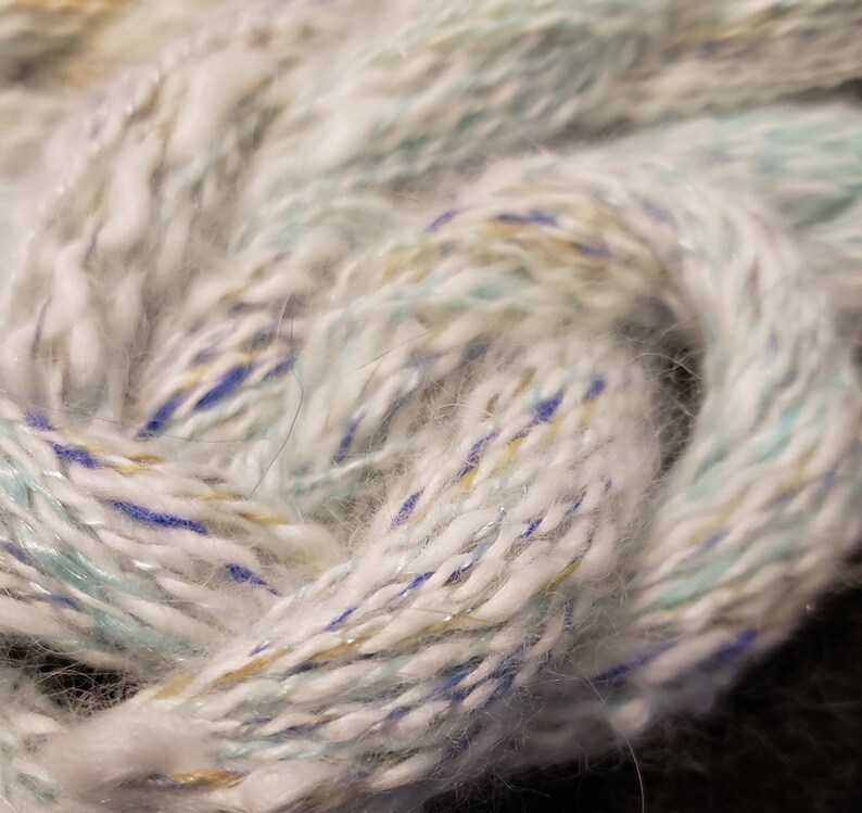 May include: White yarn with blue, green, and gold accents. The yarn is soft and fluffy, and it has a subtle shimmer.