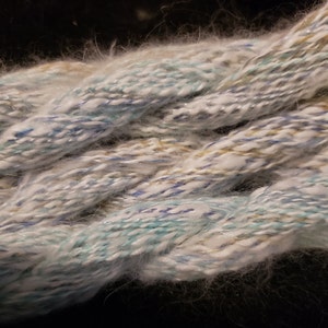 May include: A close-up of a skein of yarn with a white, blue, and gold color scheme. The yarn is soft and fluffy, with a subtle shimmer.